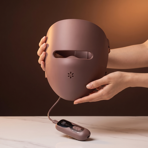 Luxéra LED mask held in two hands to show its size, with the handheld controller resting on marble