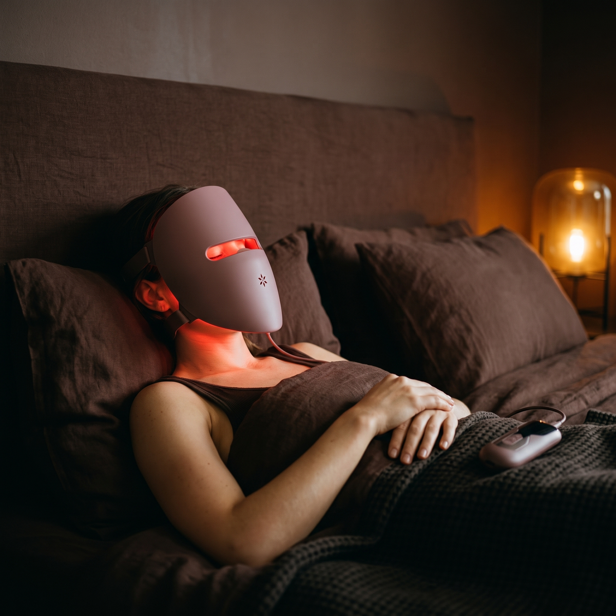 Woman resting back on linen pillows wearing the Luxéra LED mask lit in red, controller beside her